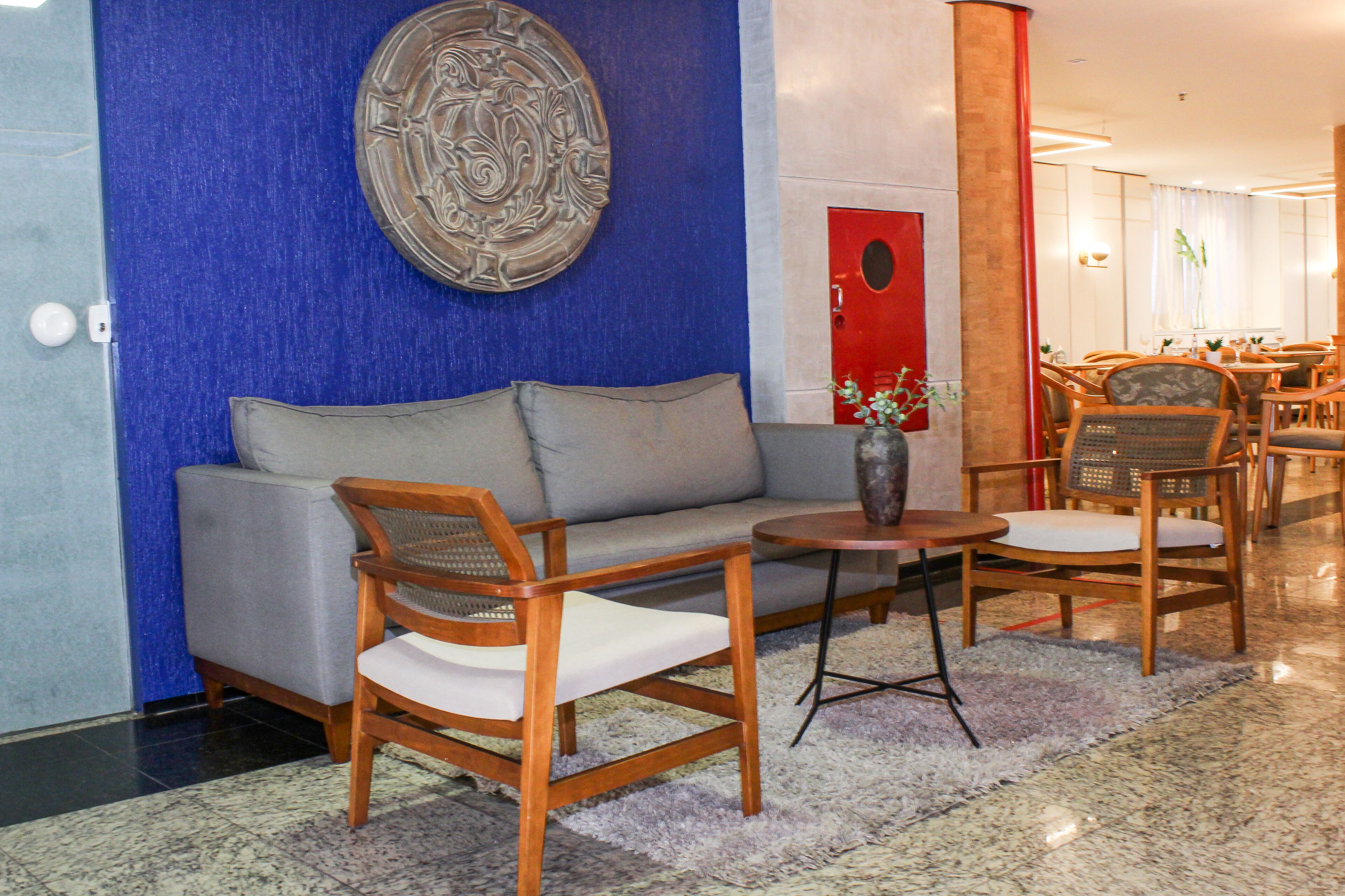 A modern lobby with a blue accent wall, a grey sofa, wooden chairs, and a small round table with a vase.