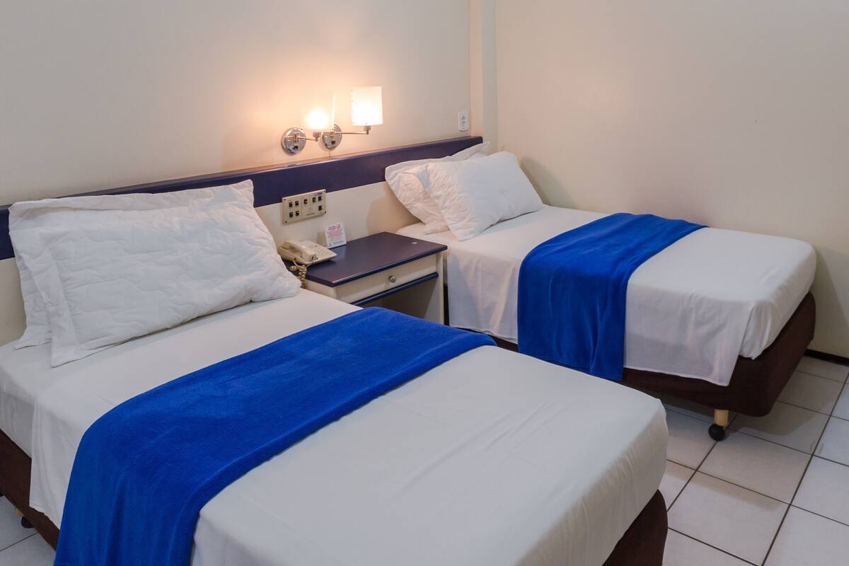 Dos camas individuales con sábanas blancas y colchas azules, una mesita de noche con un teléfono, y una lámpara de pared.
