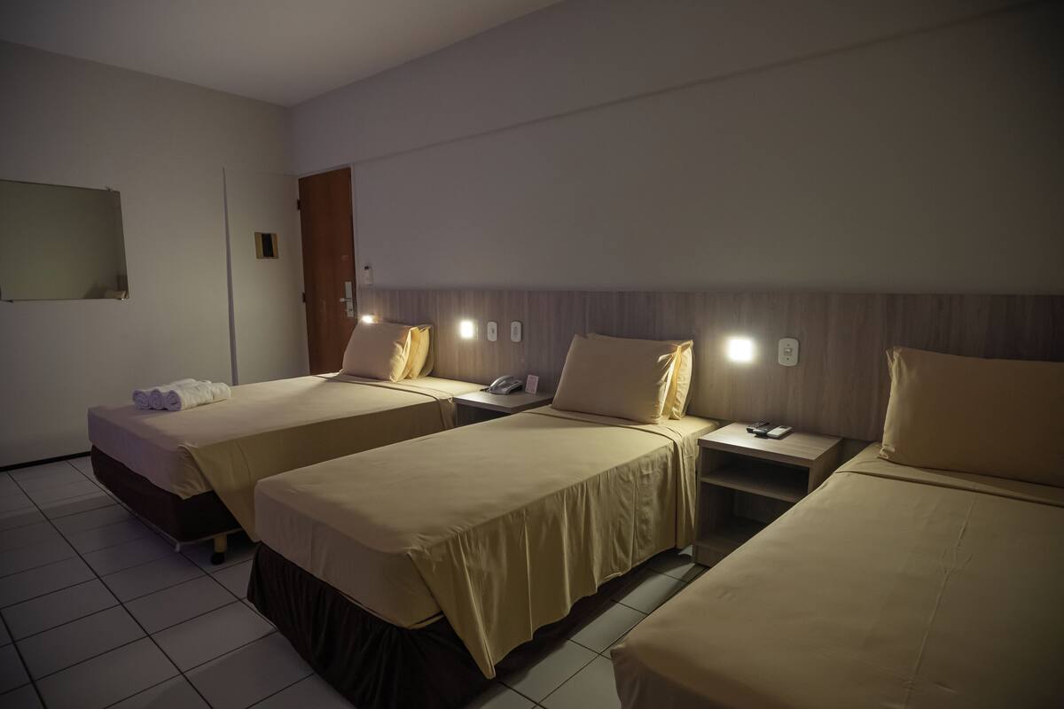 Habitación de hotel con tres camas individuales, una mesita de noche con lámpara y un espejo en la pared.