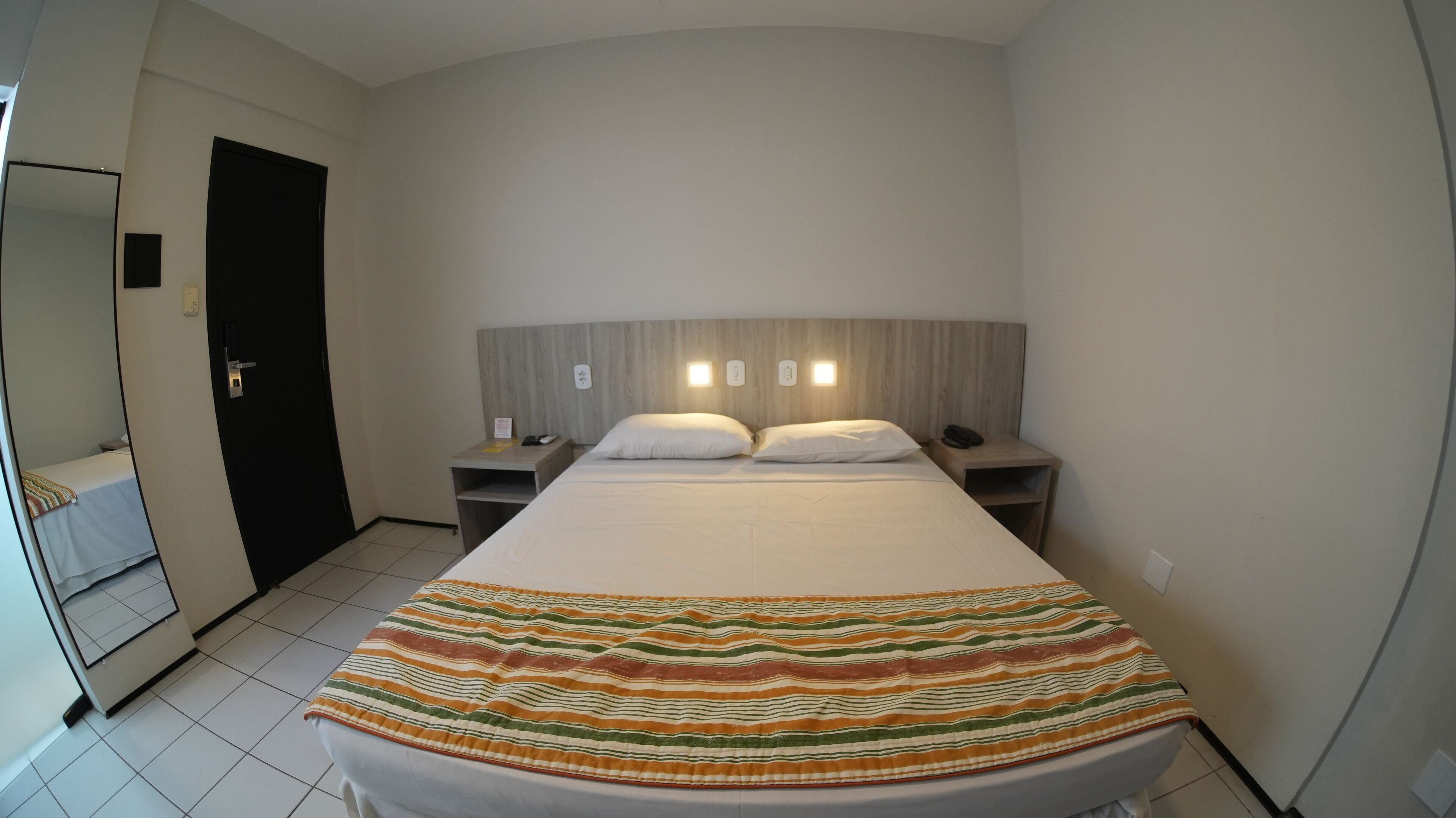 Quarto de hotel com uma cama grande, duas mesas de cabeceira, um criado-mudo, cabeceira de madeira e um espelho.