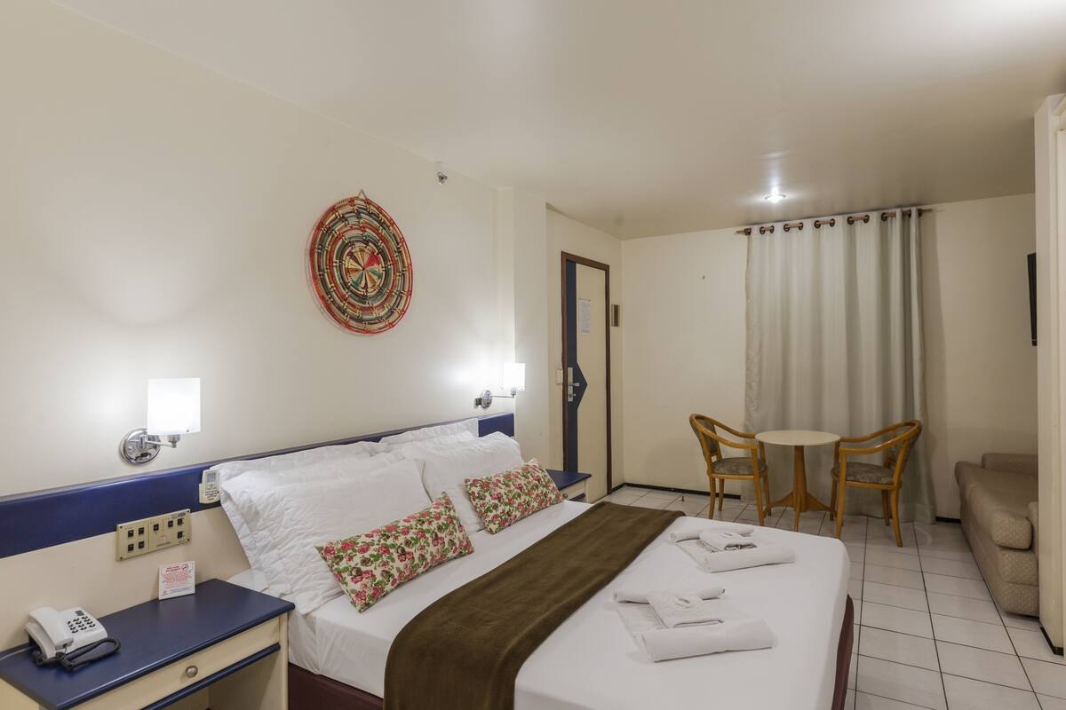 Una habitación de hotel con cama, mesita de noche, lámpara, decoración en la pared y una pequeña mesa con sillas.