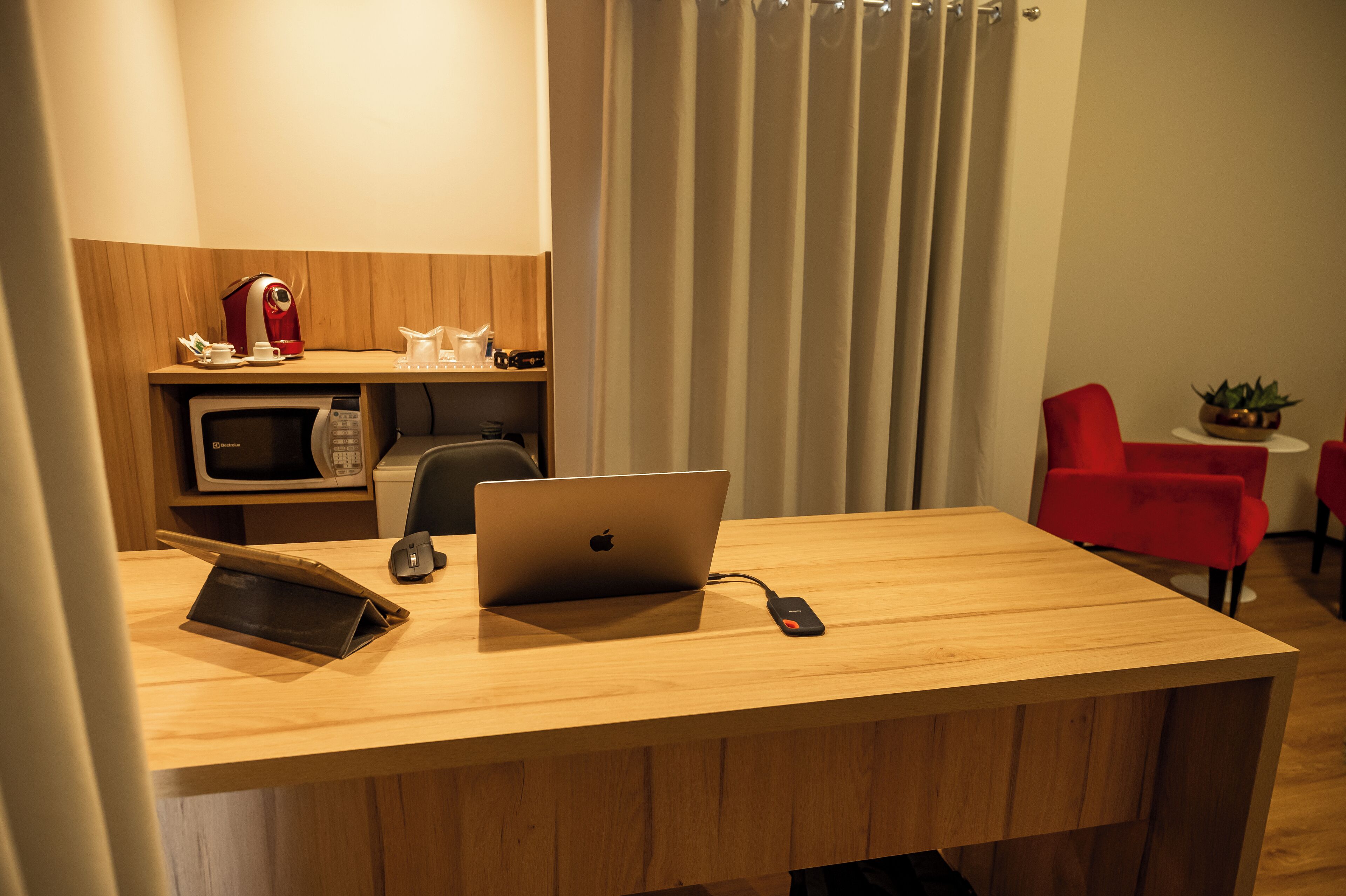 A hotel room with a wooden desk, a laptop, a microwave, a coffee maker, a red chair, and a small table with a plant.