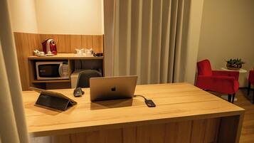 A hotel room with a wooden desk, a laptop, a microwave, a coffee maker, a red chair, and a small table with a plant.