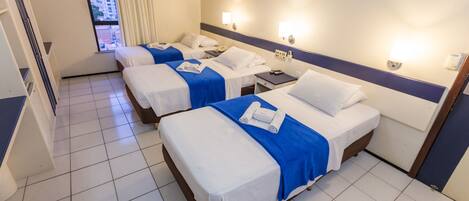 A hotel room with three single beds, each with a blue bedspread and white linens. There are bedside tables with lamps and a telephone. The room has a large window with curtains, and the floor is tiled.
