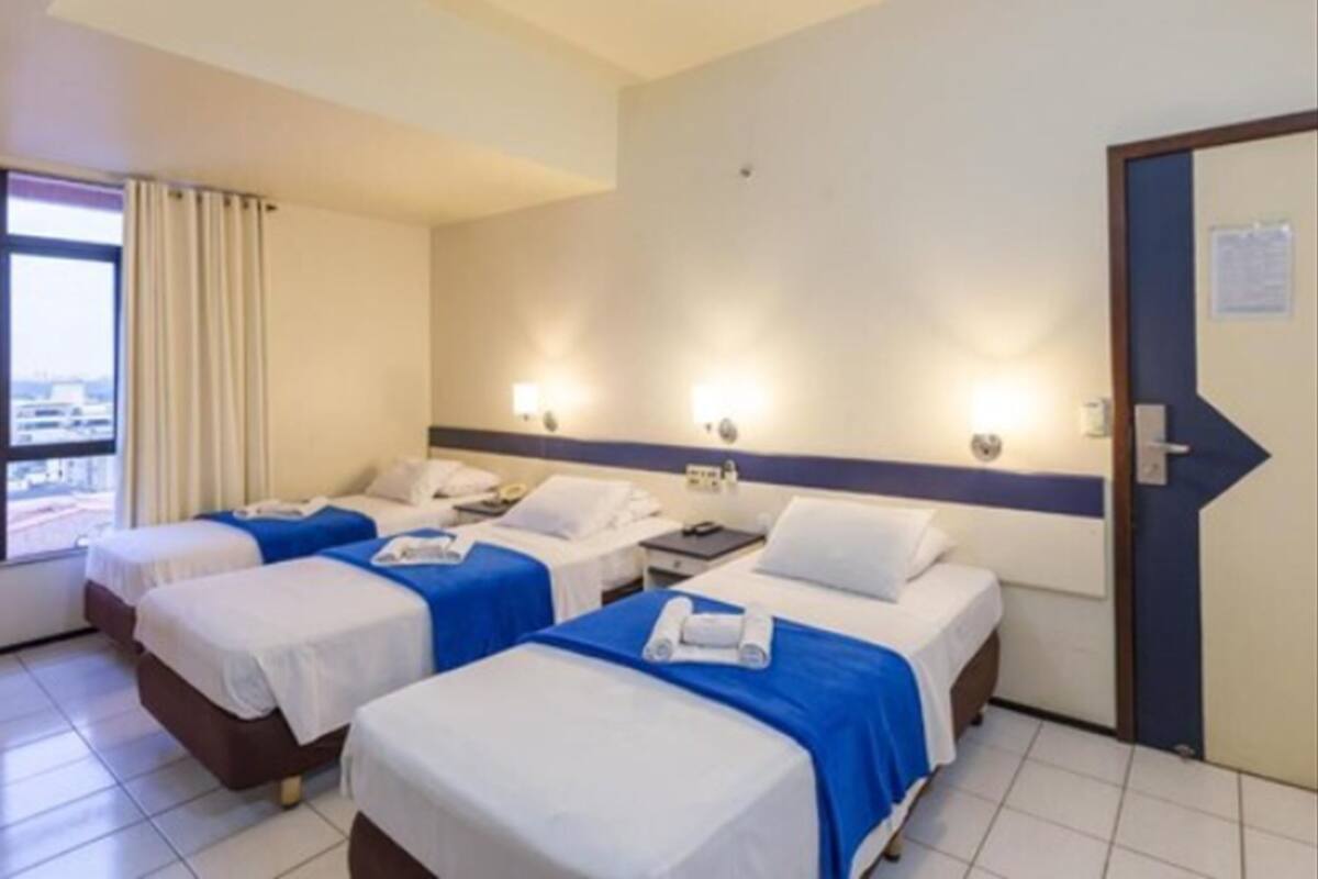 Habitación de hotel con tres camas, cada una con cubrecama azul y ropa blanca. Hay luces fijadas a la pared y una puerta con marco azul.