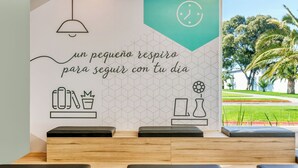 A reception area with a wall featuring a geometric pattern and a motivational quote in Spanish. There are wooden benches and a view of palm trees through a window.