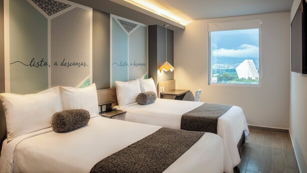 A hotel room with two beds, a large window with a city view, and a wall with decorative text.