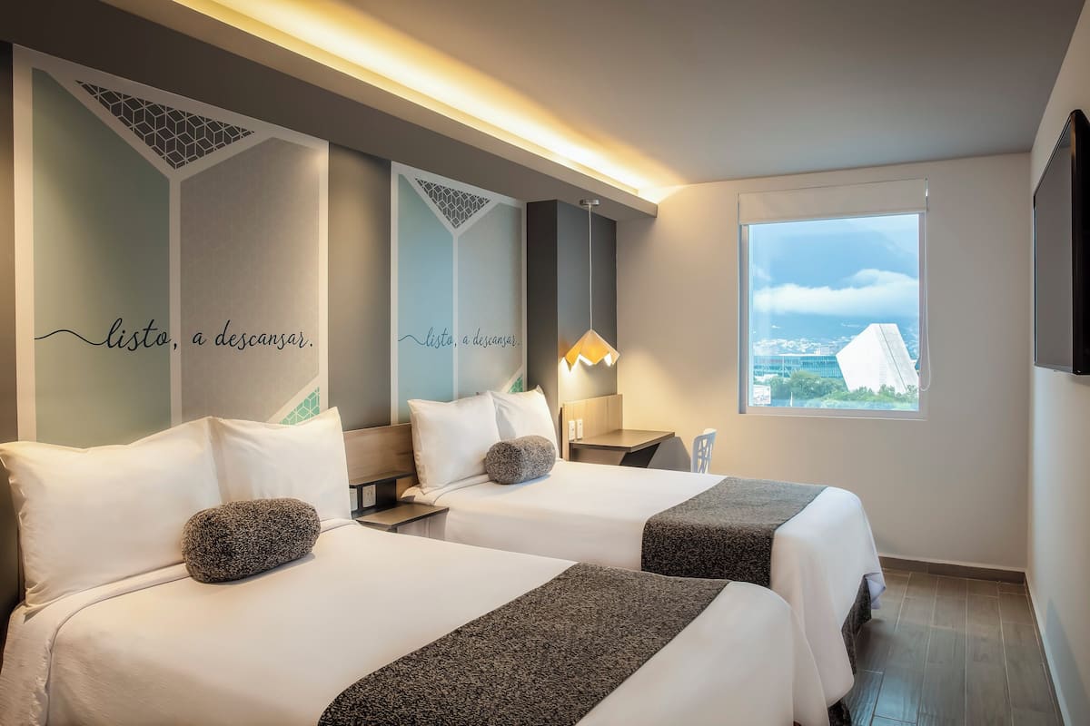 Une chambre d’hôtel avec deux lits, une grande fenêtre donnant sur la ville et un mur orné d’un motif décoratif.