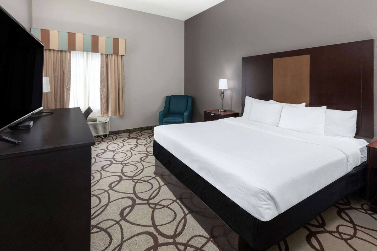 Ein Hotelzimmer mit einem großen Bett, einem Schreibtisch mit Flachbildfernseher, einem Sessel, einem Nachttisch mit Lampe und einem Fenster mit Vorhängen.