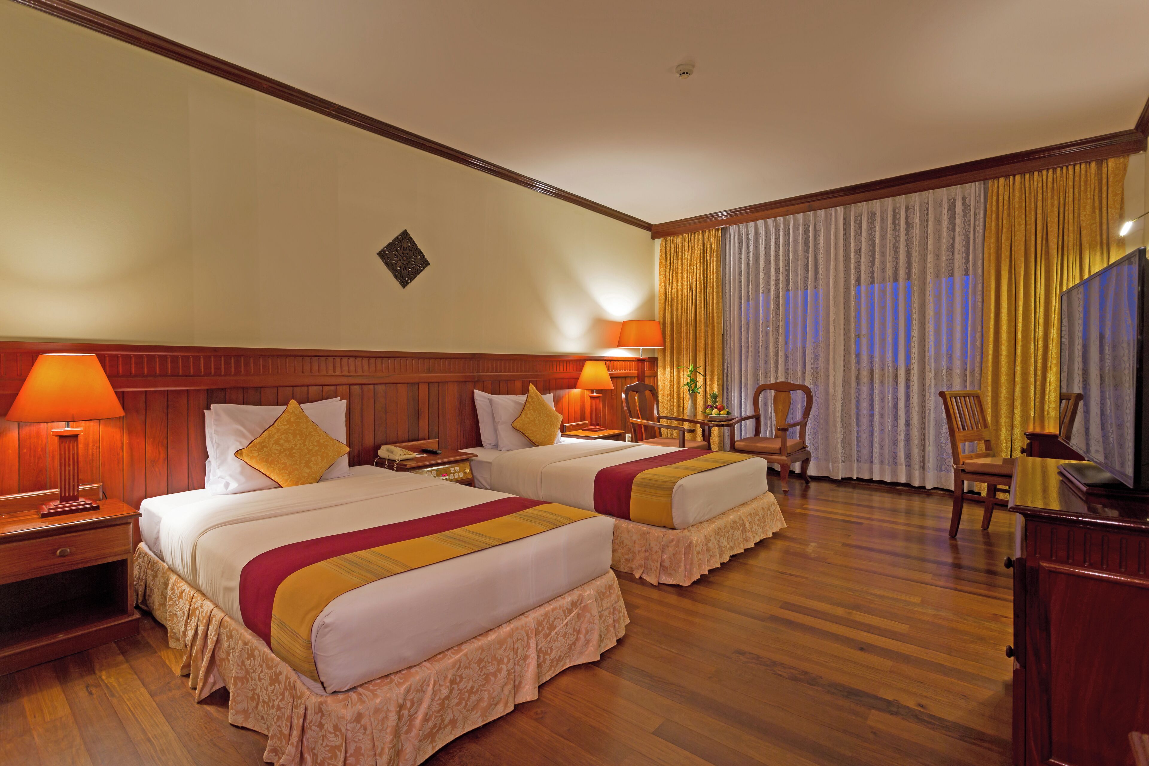 A hotel room with two beds, wooden flooring, a desk with a lamp, a chair, and a large window with curtains.
