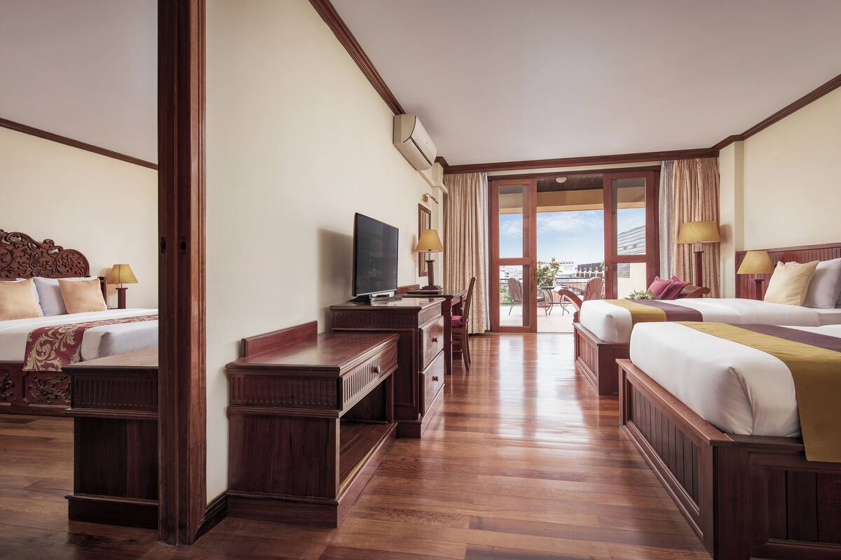 A hotel room with two beds, a TV, wooden furniture, and a view of the city through large windows.