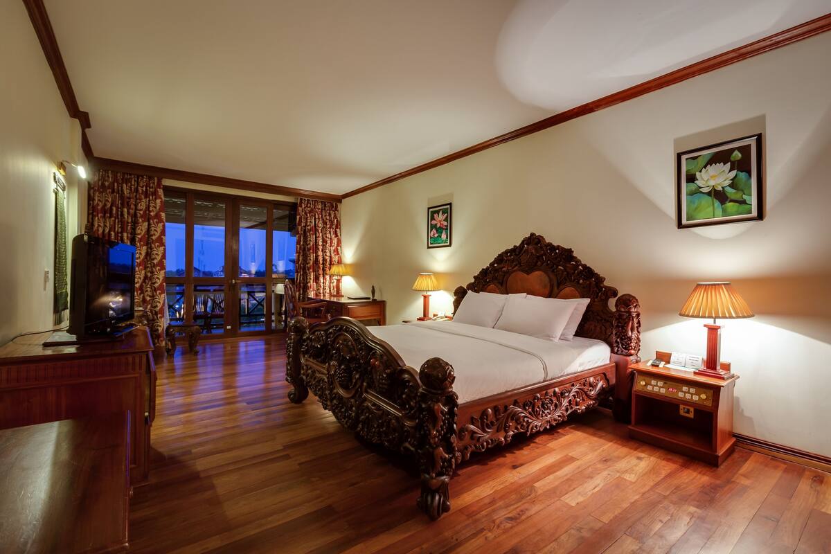 A spacious bedroom with a large wooden bed, a television, and a nightstand with a lamp.