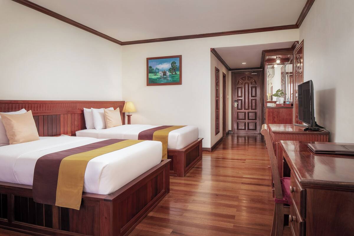 A hotel room with two beds, wooden headboards, a painting on the wall, a TV, and a desk with drawers.