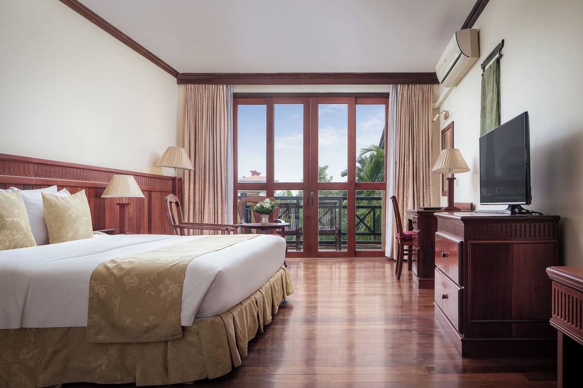A hotel room with a large bed, a TV, a desk with a chair, and a balcony with curtains.