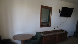 A room with a round table, a green chair, a wooden dresser, a mirror, and a flat-screen TV mounted on the wall. - Jim Butler Inn & Suites (Tonopah)