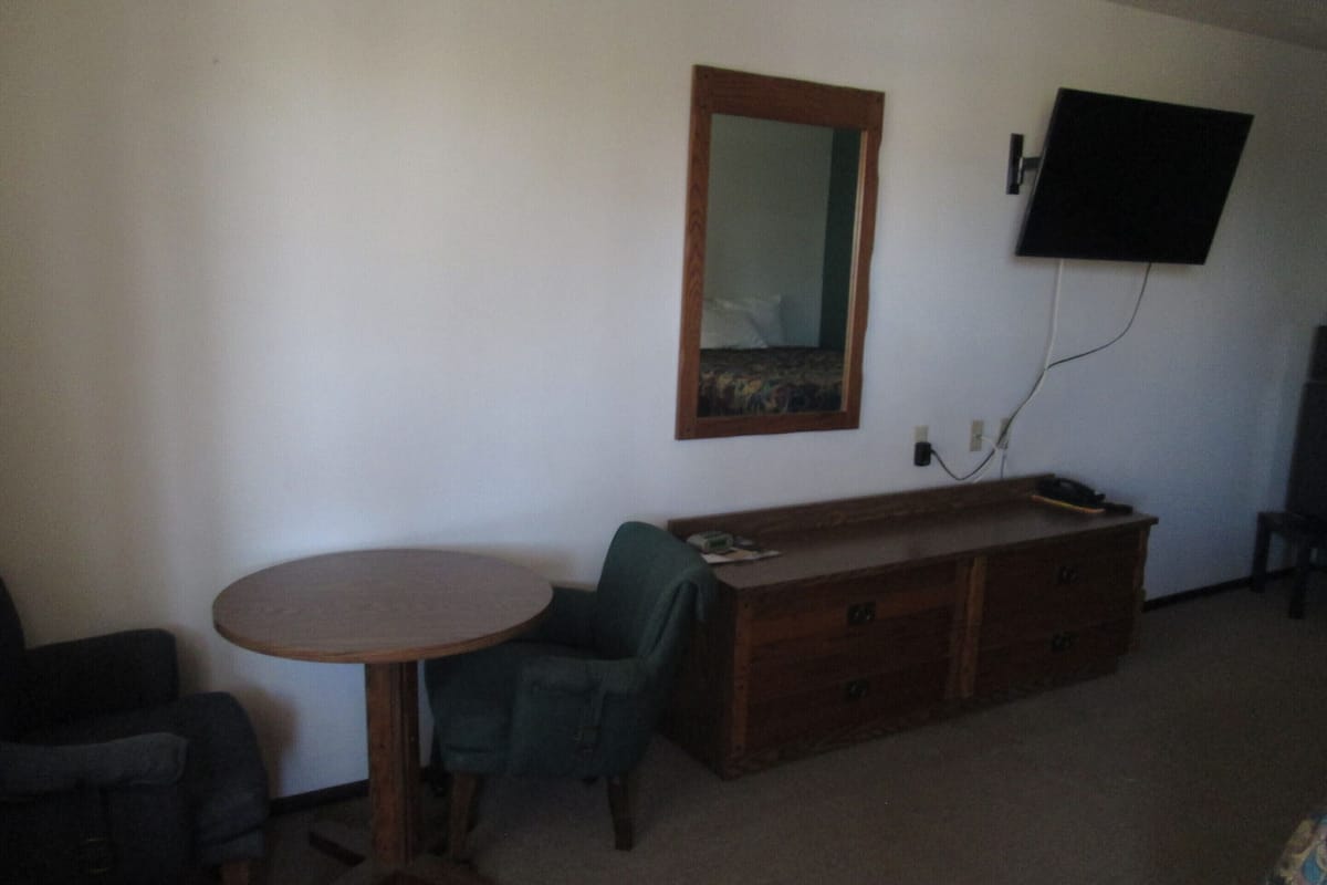 A room with a round table, a green chair, a wooden dresser, a mirror, and a flat-screen TV mounted on the wall.