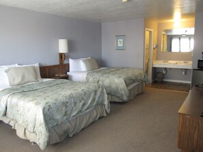 A hotel room with two beds, a nightstand, a lamp, a bathroom with a sink and mirror, and a wooden desk.