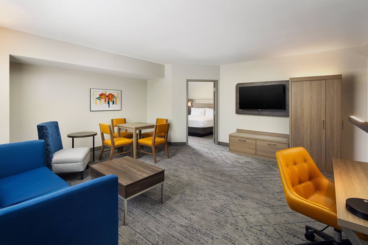 Hotelkamer met een blauwe bank, gele stoelen, een houten salontafel, een aan de muur bevestigde televisie en een bed met een wit hoofdeinde.