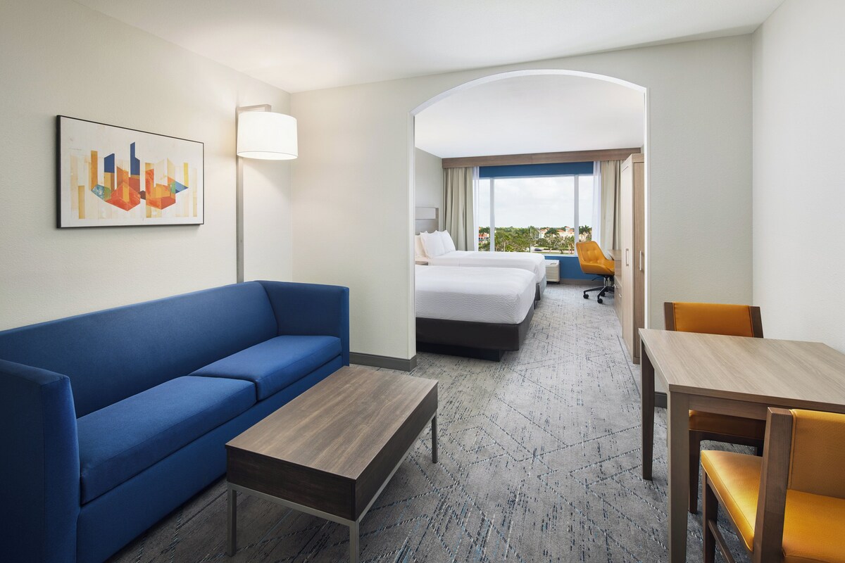 Une chambre d’hôtel avec un canapé bleu, une table basse en bois, une table à manger en bois avec des chaises jaunes, un lit avec une literie blanche et une vue sur la ville par la fenêtre.