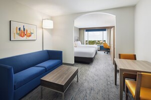Une chambre d’hôtel avec un canapé bleu, une table basse en bois, un lit avec une literie blanche, un bureau avec une chaise et une grande fenêtre donnant sur un paysage.