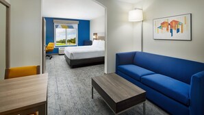 Habitación de hotel con cama, sofá azul, mesa de centro de madera y escritorio. Se ve un ventanal con vistas a zonas verdes.