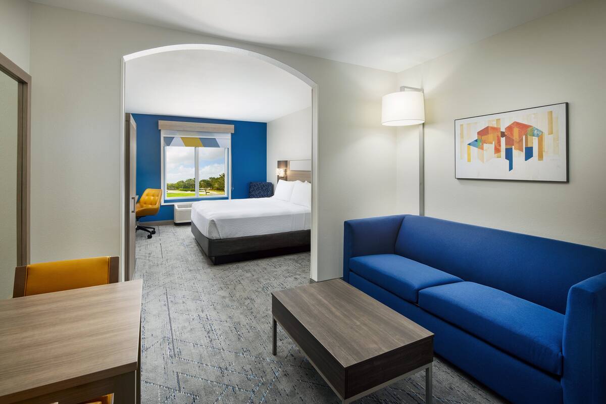 Een hotelkamer met een bed, een blauwe bank, een houten salontafel en een bureau. Er is een raam met uitzicht op groen.