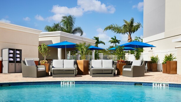 Zona junto a la piscina con sillones de descanso y sombrillas, una piscina azul cristalina y una valla blanca.