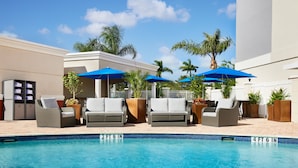 Zona junto a la piscina con sillones de descanso y sombrillas, una piscina azul cristalina y una valla blanca.