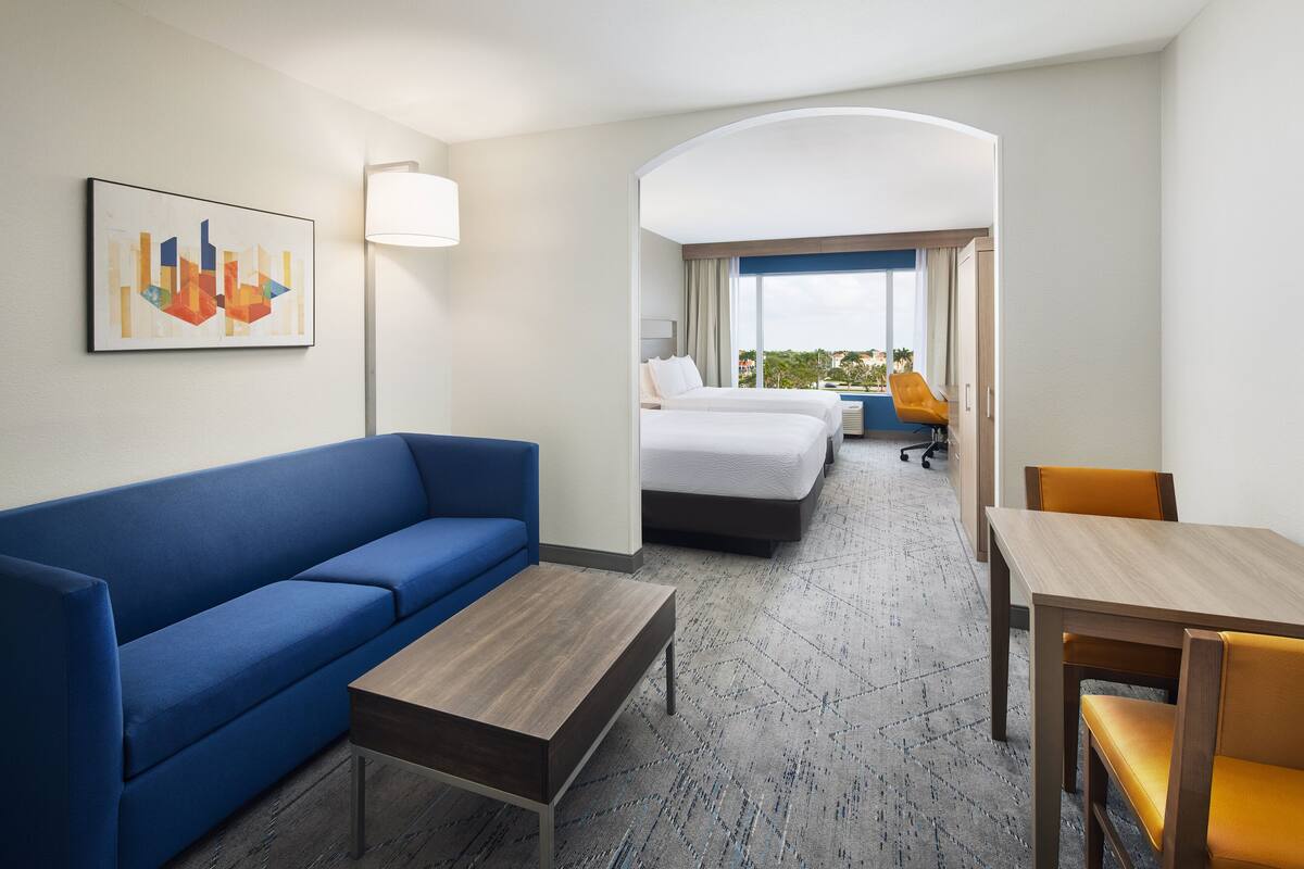 Hotelkamer met een blauwe bank, een houten bijzettafeltje, een houten eettafel met gele stoelen, een bed met witte beddengoed en uitzicht op de stad door een raam.