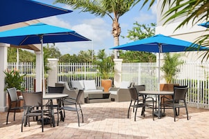 An outdoor patio area with round tables and chairs, blue umbrellas, and a white fence.
