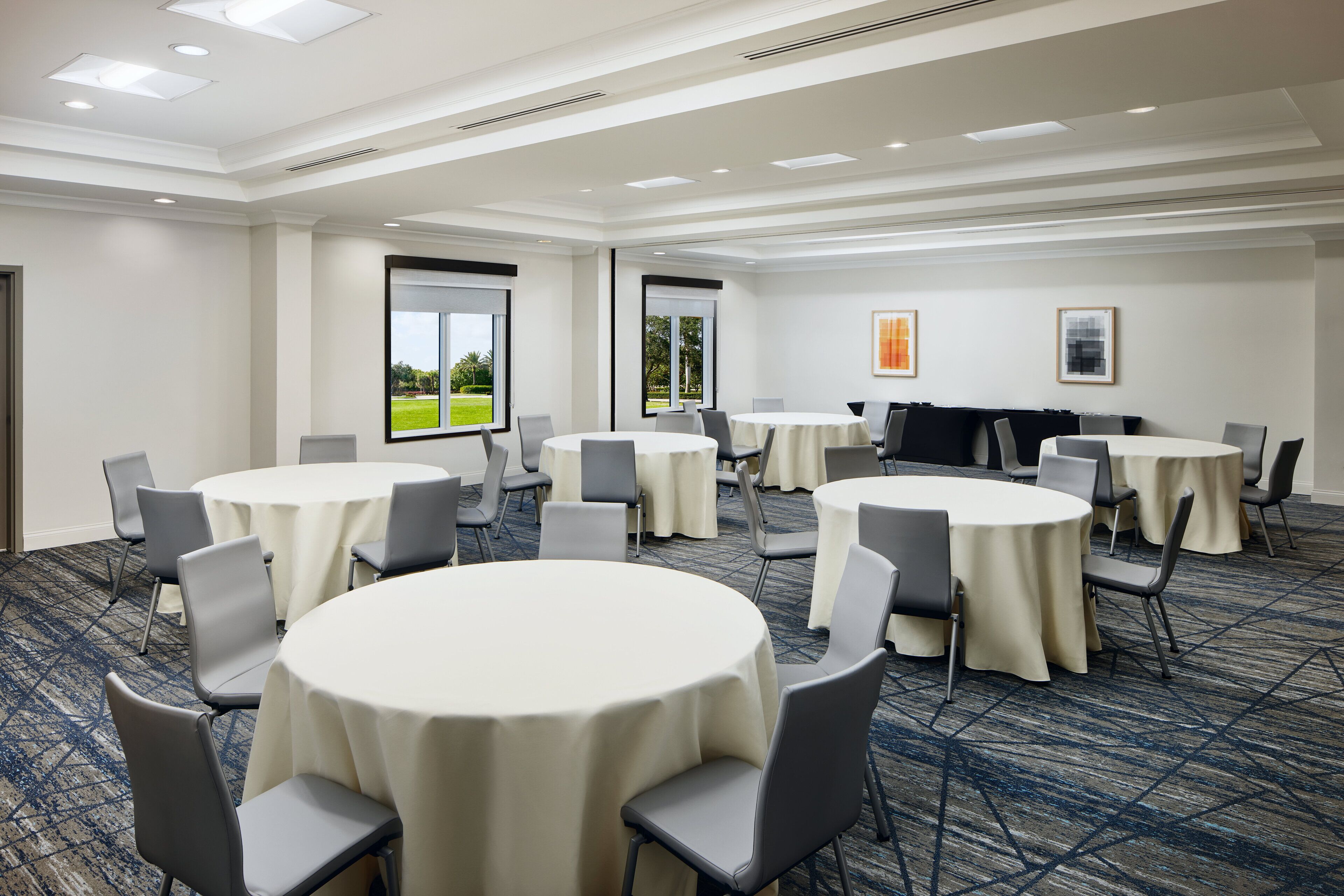 A conference room with multiple round tables and chairs, large windows with a view of greenery, and framed artwork on the walls.