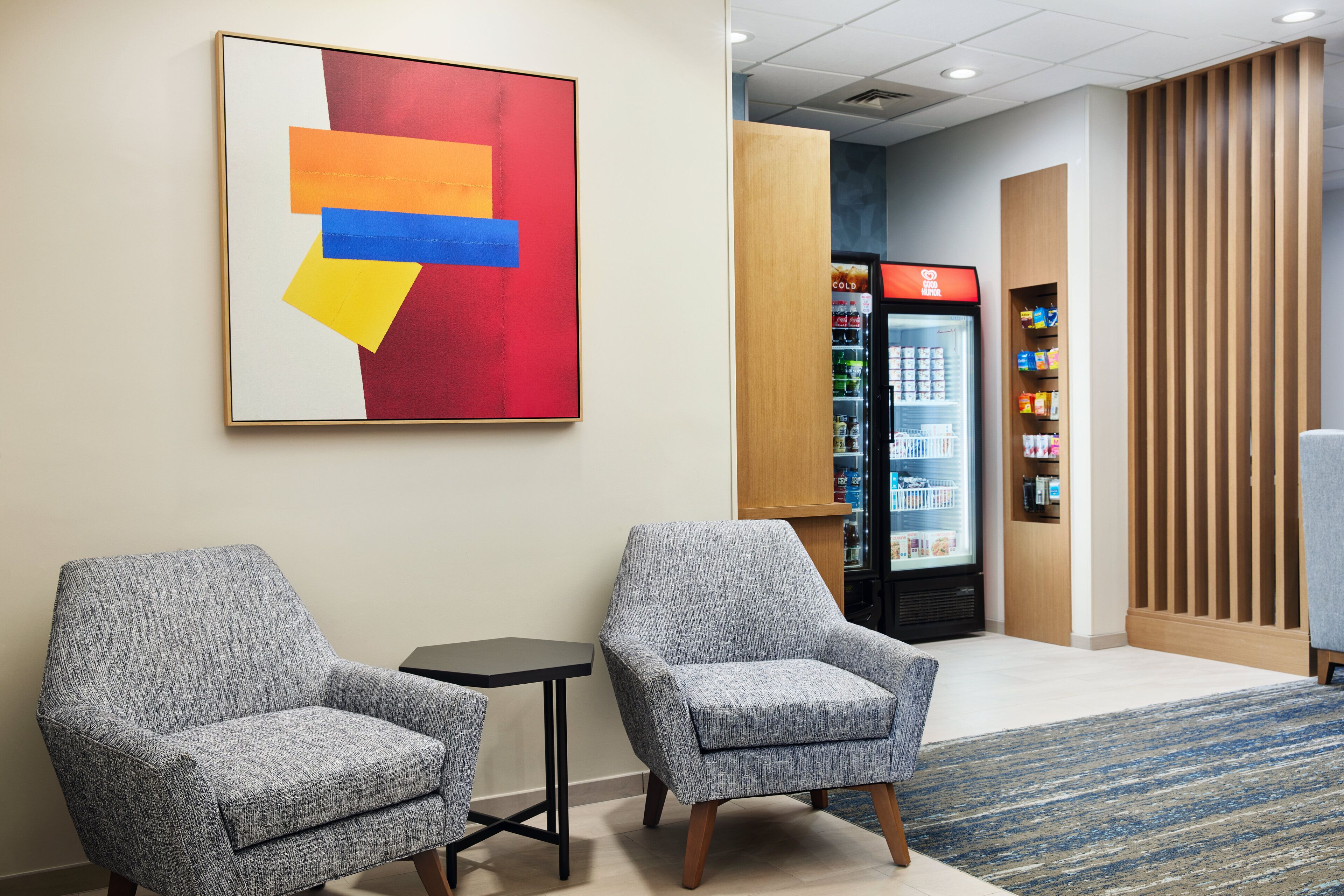 A hotel lobby with two grey armchairs, a small table, a colorful abstract painting, a refrigerator, and a wooden partition.