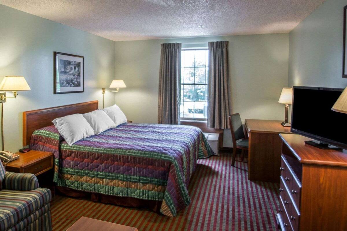 Una habitación de hotel con cama, escritorio, silla, televisor y ventana con cortinas.