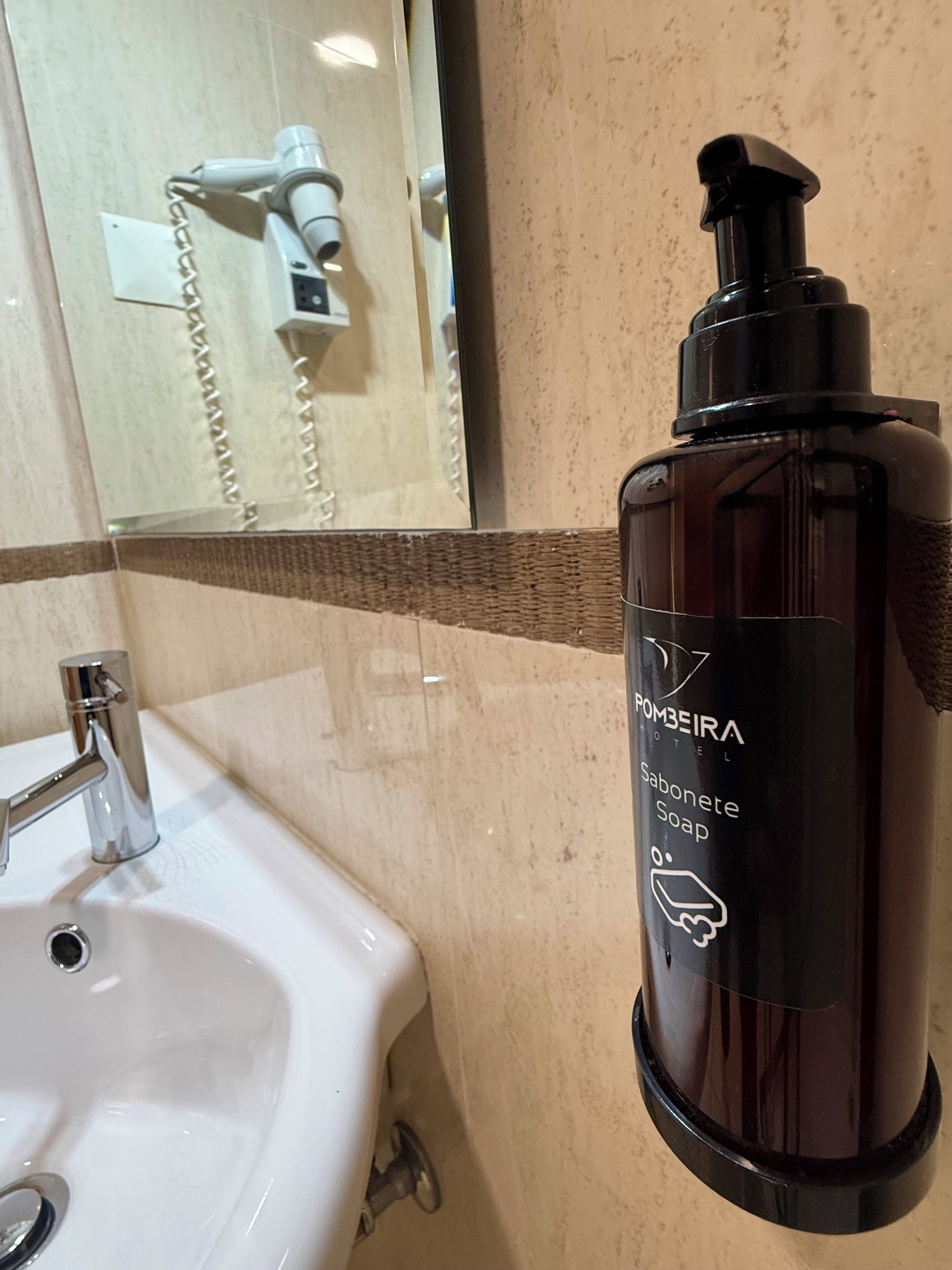 Een badkamer met een witte wastafel, een spiegel en een fles zeep met de tekst 'POMBEIRA HOTEL'.