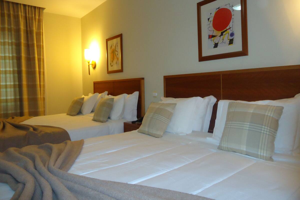 A hotel room with two beds, a large window with curtains, a wall-mounted lamp, and a framed picture.