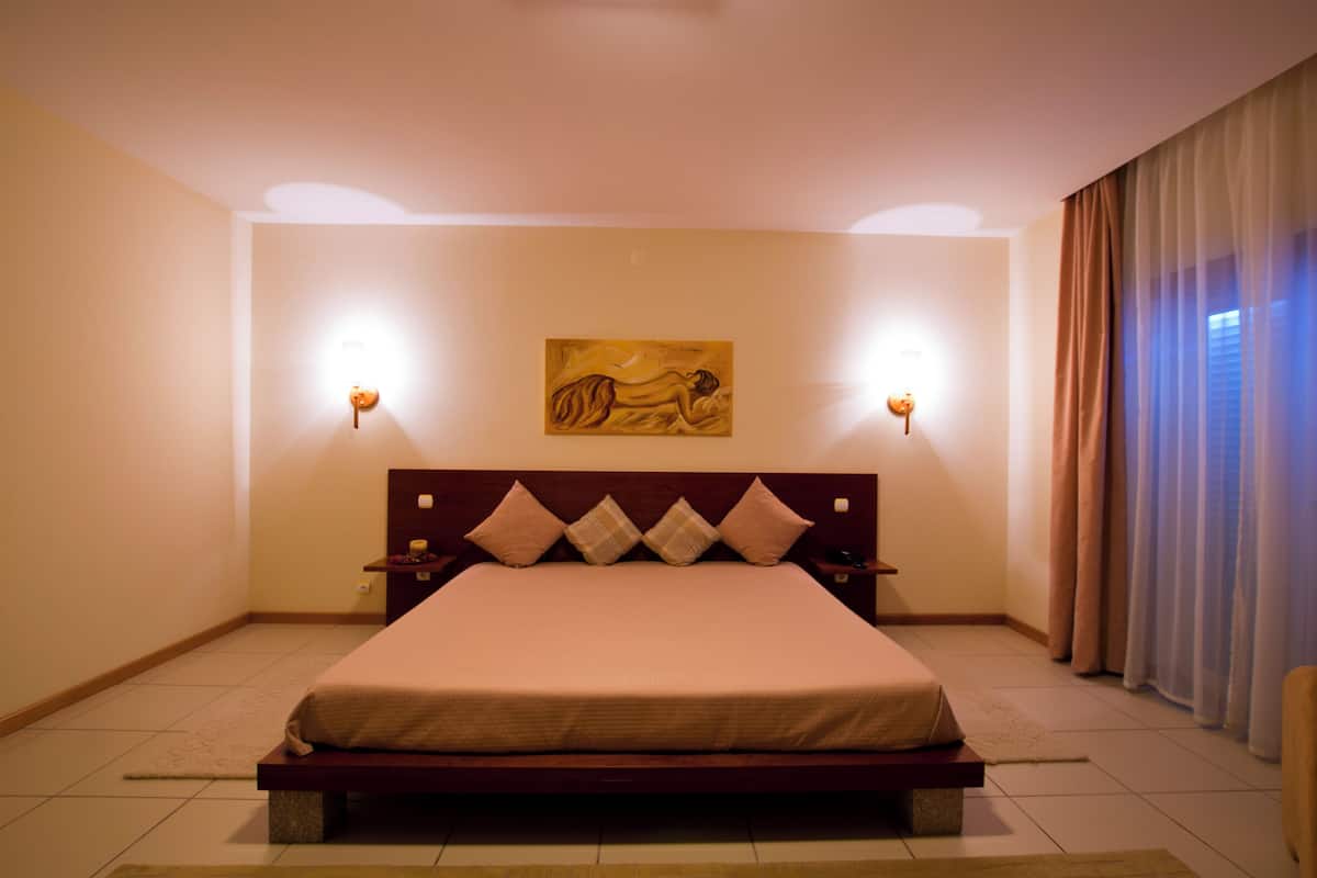 A bedroom with a large bed, two bedside tables, a wall-mounted artwork, and a window with curtains.