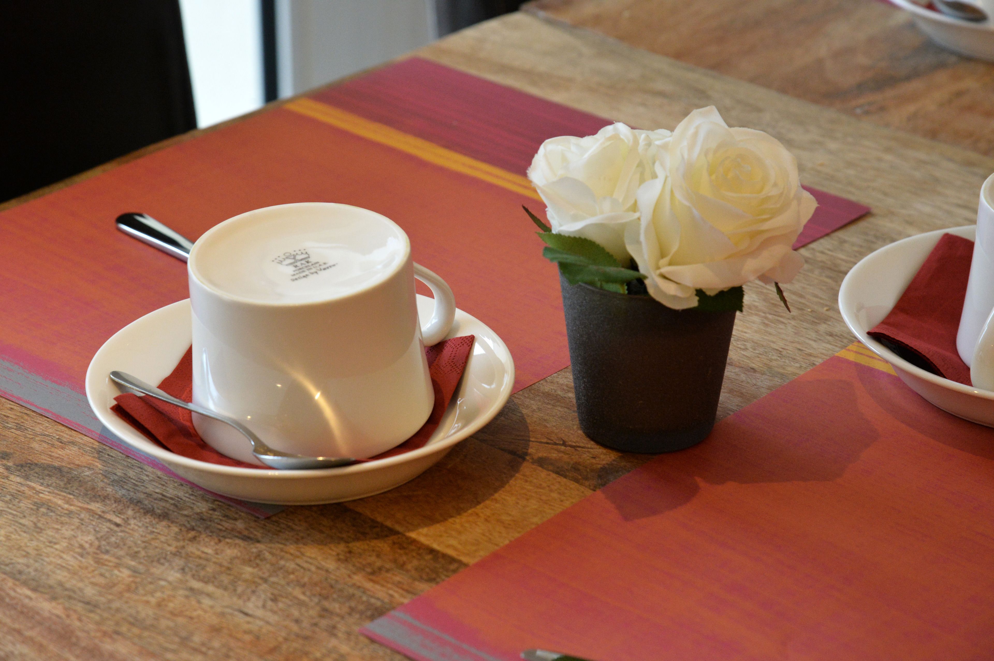 A table with a white cup and saucer, a spoon, and a small vase with white flowers.