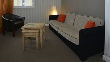 Un salon comprenant un fauteuil en cuir noir, une table basse en bois, un canapé gris avec des coussins orange et une lampe.