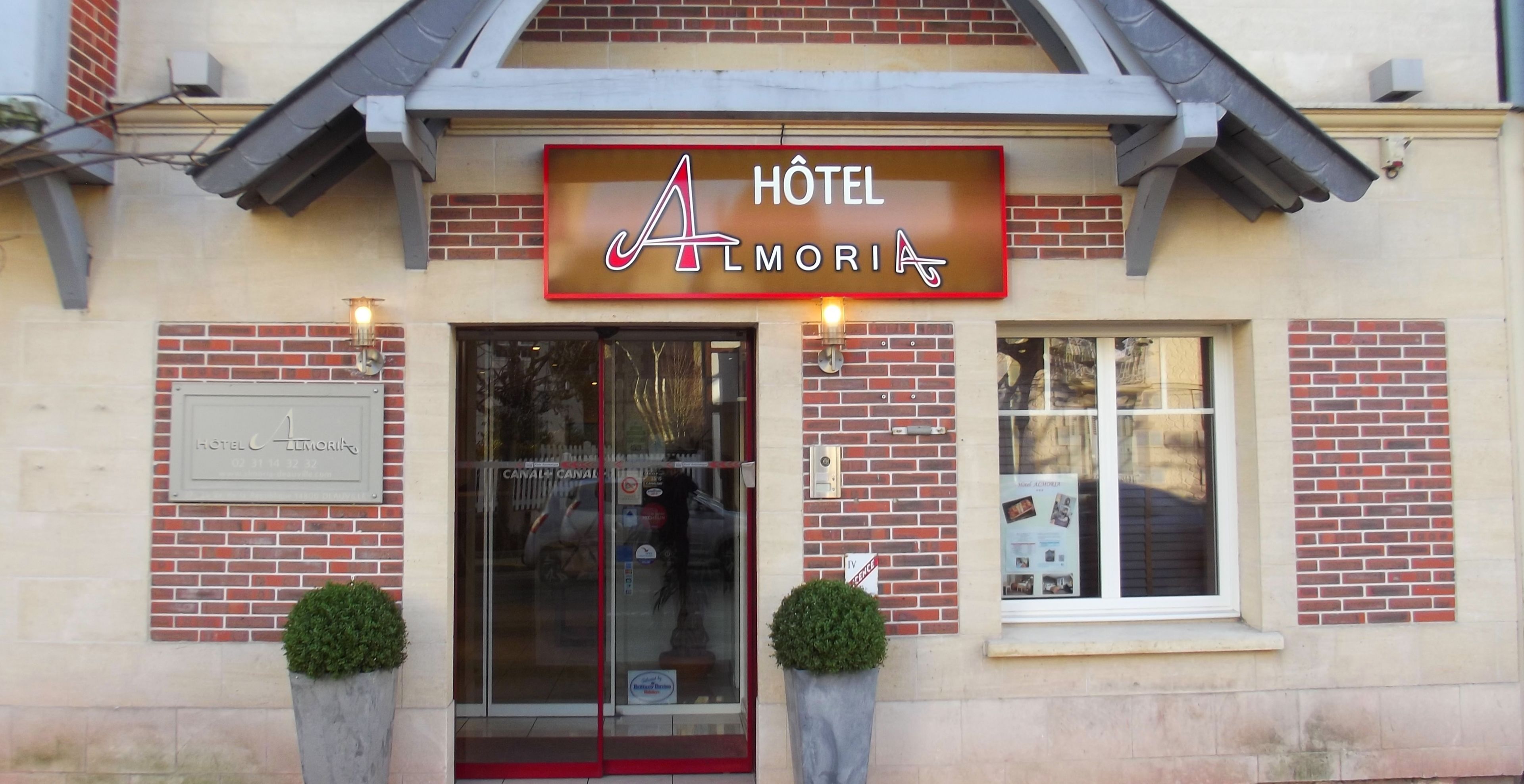 L'esterno dell'Hotel Almoria, con un cartello ben visibile, un ingresso in vetro e due piante in vaso.