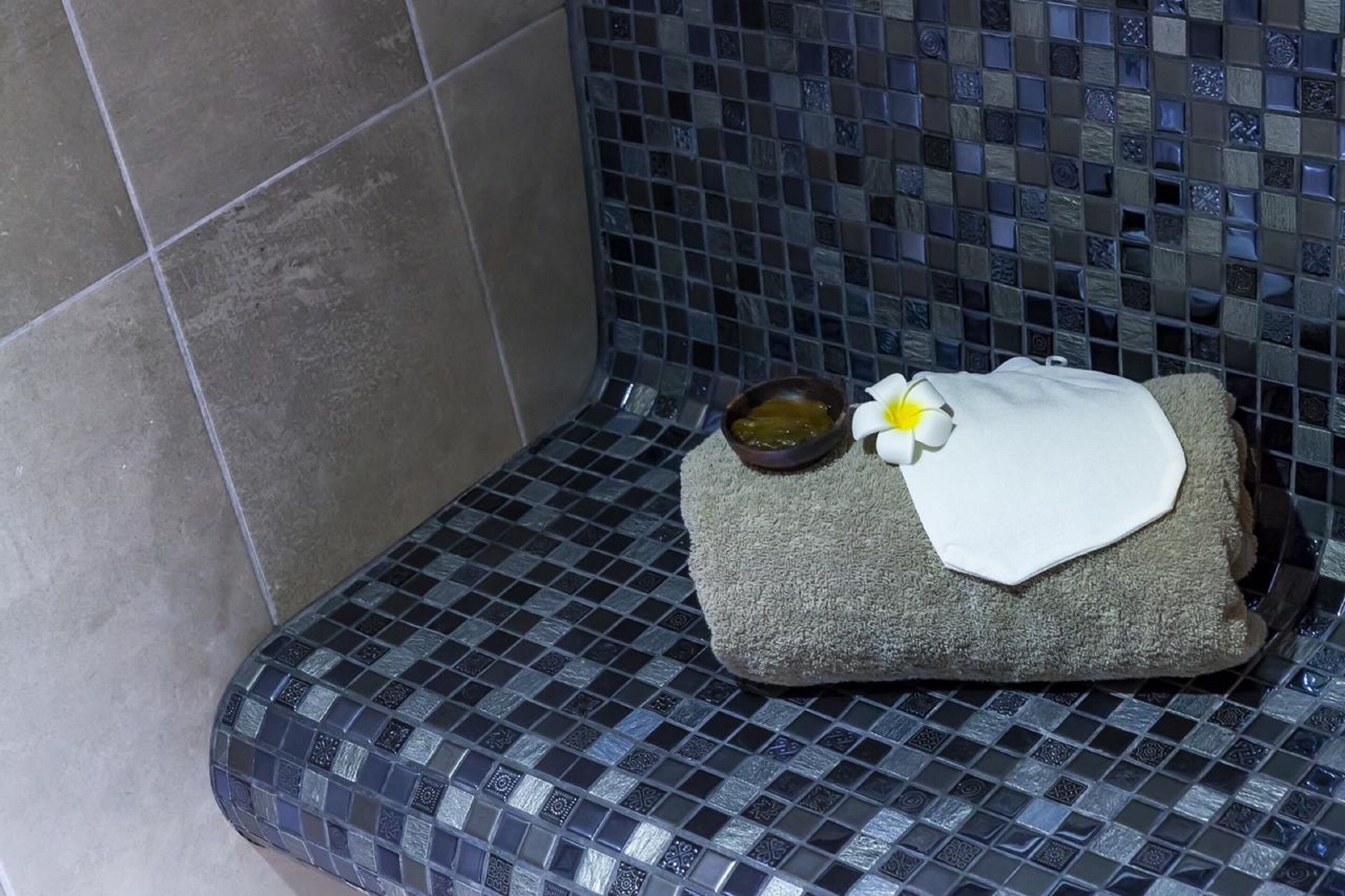 A bathroom with a tiled bench, a small bowl of oil, and a white towel with a flower.