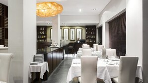 Un comedor moderno con mesas y sillas blancas, un área de bar central con una vitrina de vinos, y una lámpara colgante grande.