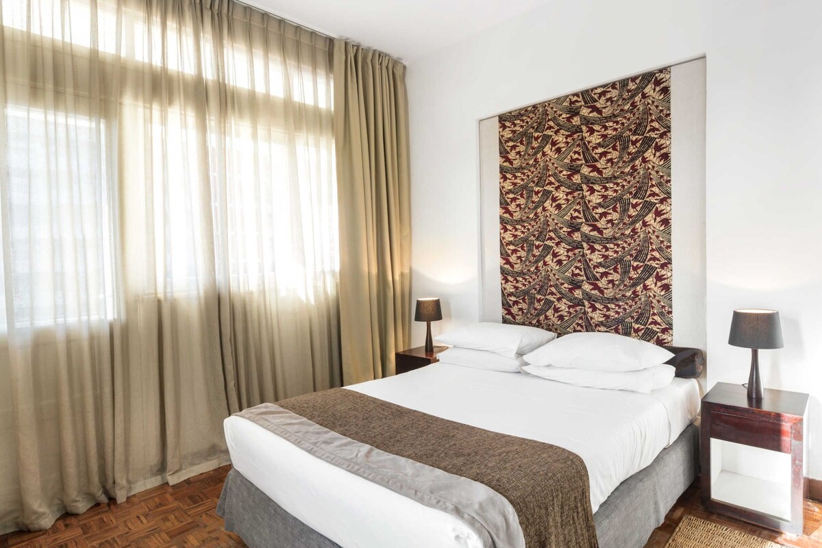 A hotel room with a large bed, a patterned headboard, two bedside tables with lamps, and a window with sheer curtains.