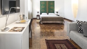 Una habitación de hotel moderna con una televisión de pantalla plana, un escritorio con lámpara, una cama con cabecera estampada en verde, un sofá gris y una lámpara colgante grande.