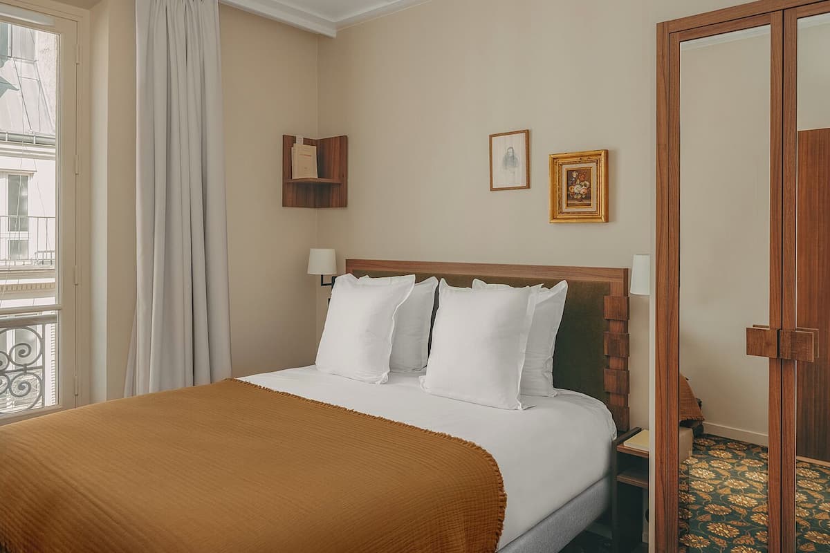 A hotel room with a bed, a nightstand, a wooden headboard, a window with curtains, and a door.
