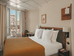 Una habitación de hotel con una cama grande, cabecera de madera, mesita de noche con lámpara, un cuadro en la pared y un balcón con mesa y sillas.