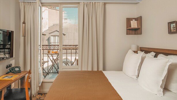 Een hotelkamer met een bed, een bureau met telefoon, een stoel, een televisie en een balkon met tafel en stoelen.