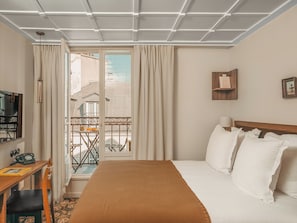 Habitación de hotel con cama, escritorio con teléfono, silla, televisión y balcón con mesa y sillas.