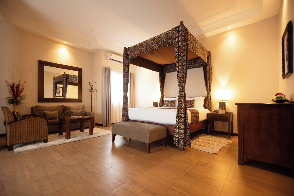 A four-poster bed with a patterned canopy, a sofa, a small table, and a bench in a well-lit room with wooden flooring.