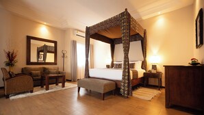 A four-poster bed with a patterned canopy, a sofa, a small table, and a bench in a well-lit room with wooden flooring.