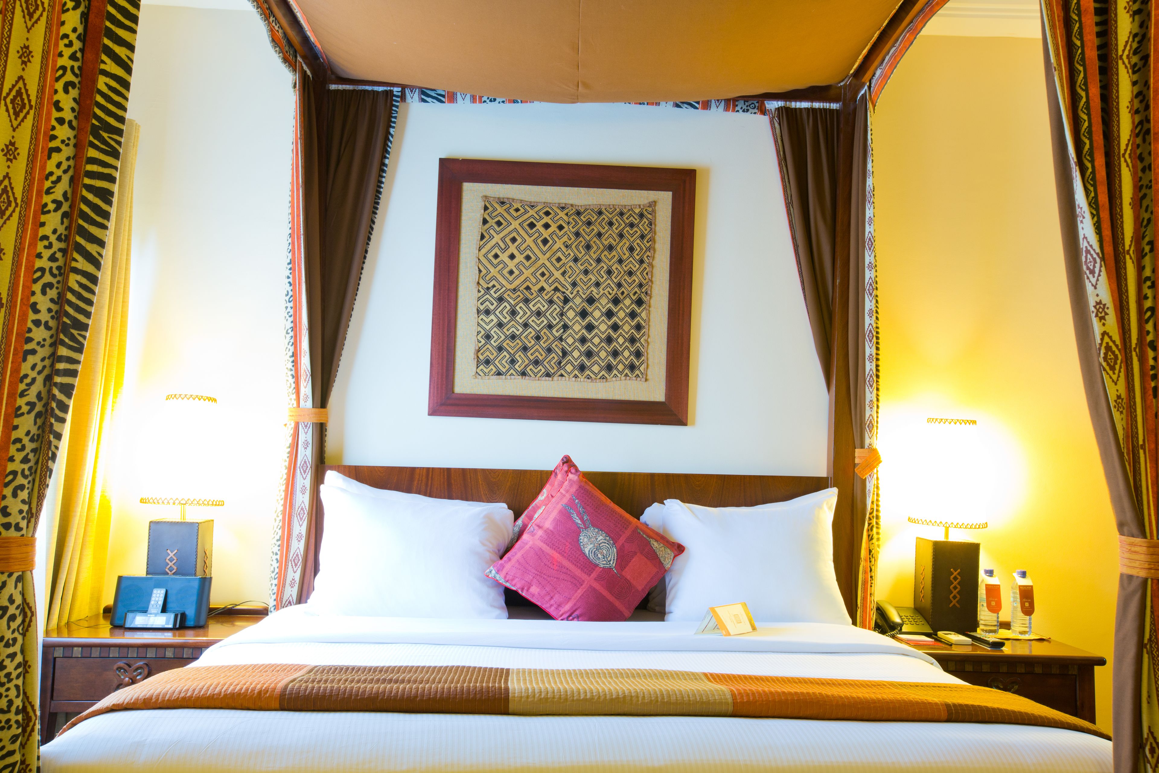 Un lit à baldaquin avec une tête de lit à motifs, un coussin décoratif rose et une table de chevet avec une lampe et un téléphone.